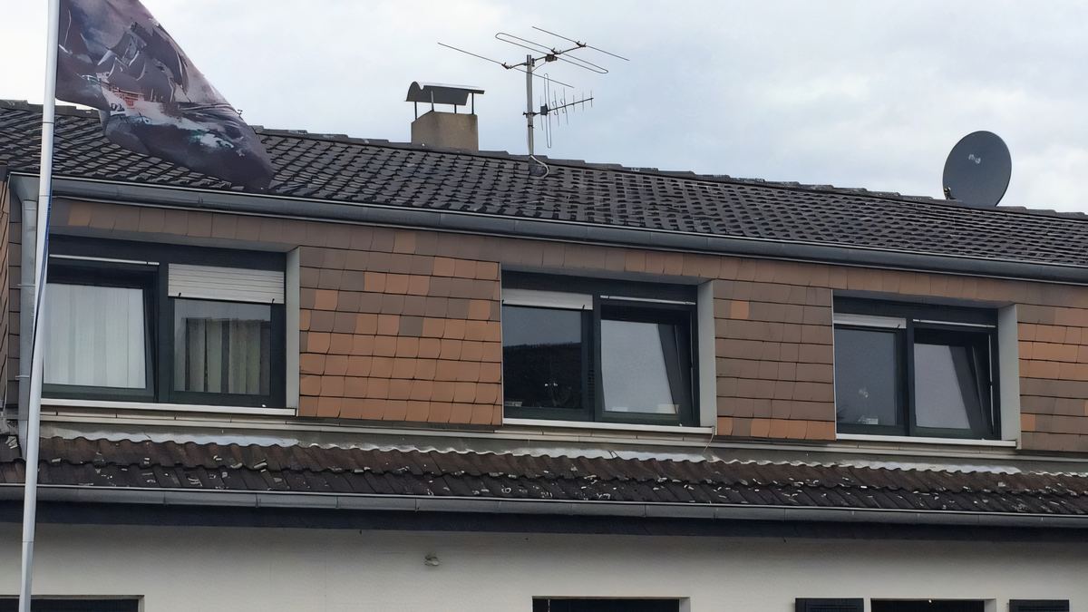Lange Gaube mit brauner Schindelbekleidung auf einem Reihenhaus, auf dem Dach Schornstein und Antenne