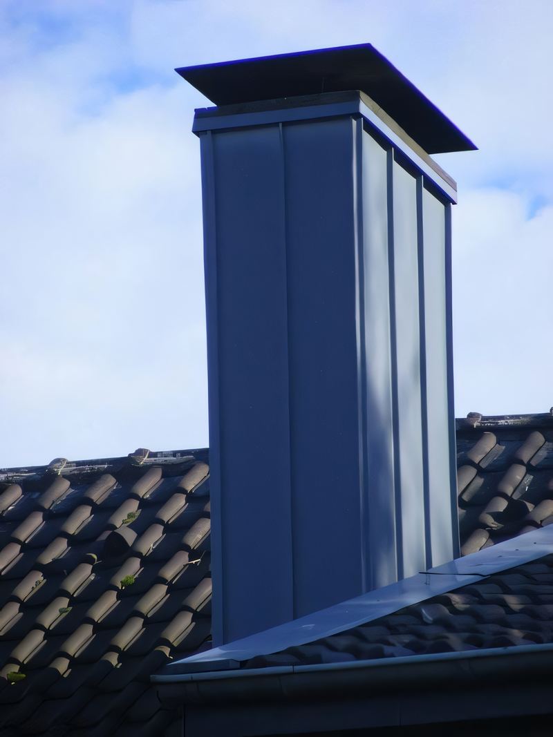 Hoher Schornstein mit blaugrauer senkrechter Metallbekleidung und Abdeckplatte auf einem dunklen Ziegeldach