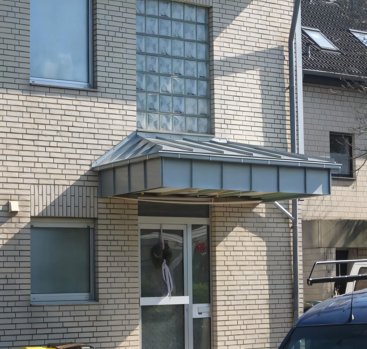 Vordach über einem Hauseingang mit grauer Metall-Stehfalzbekleidung, Klinkerfassade mit Glasbausteinen