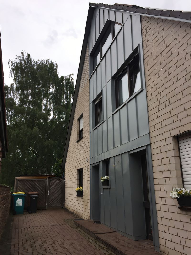 Schmales Haus mit grauer Metall-Stehfalzfassade neben hellem Klinker