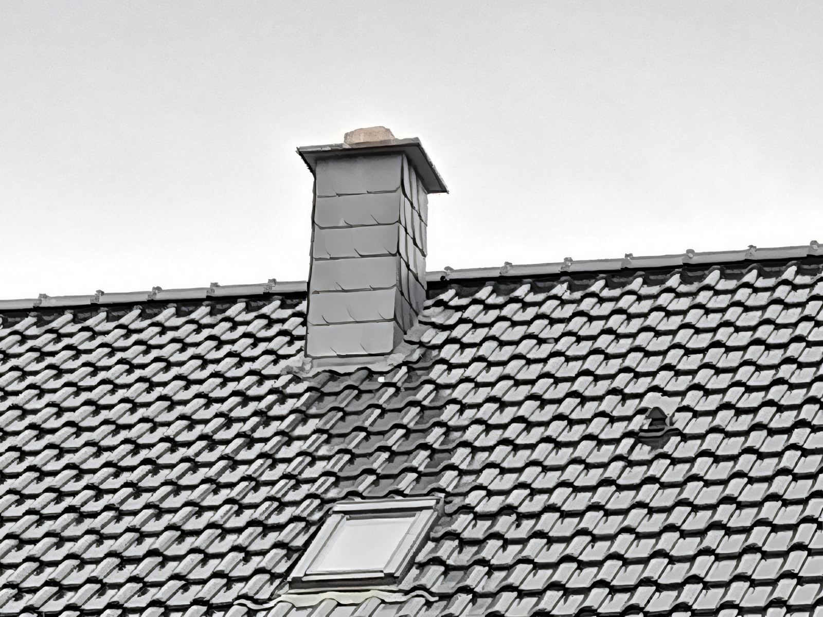 Schornstein mit grauer Schieferbekleidung auf einem grauen Ziegeldach, darunter ein Dachfenster