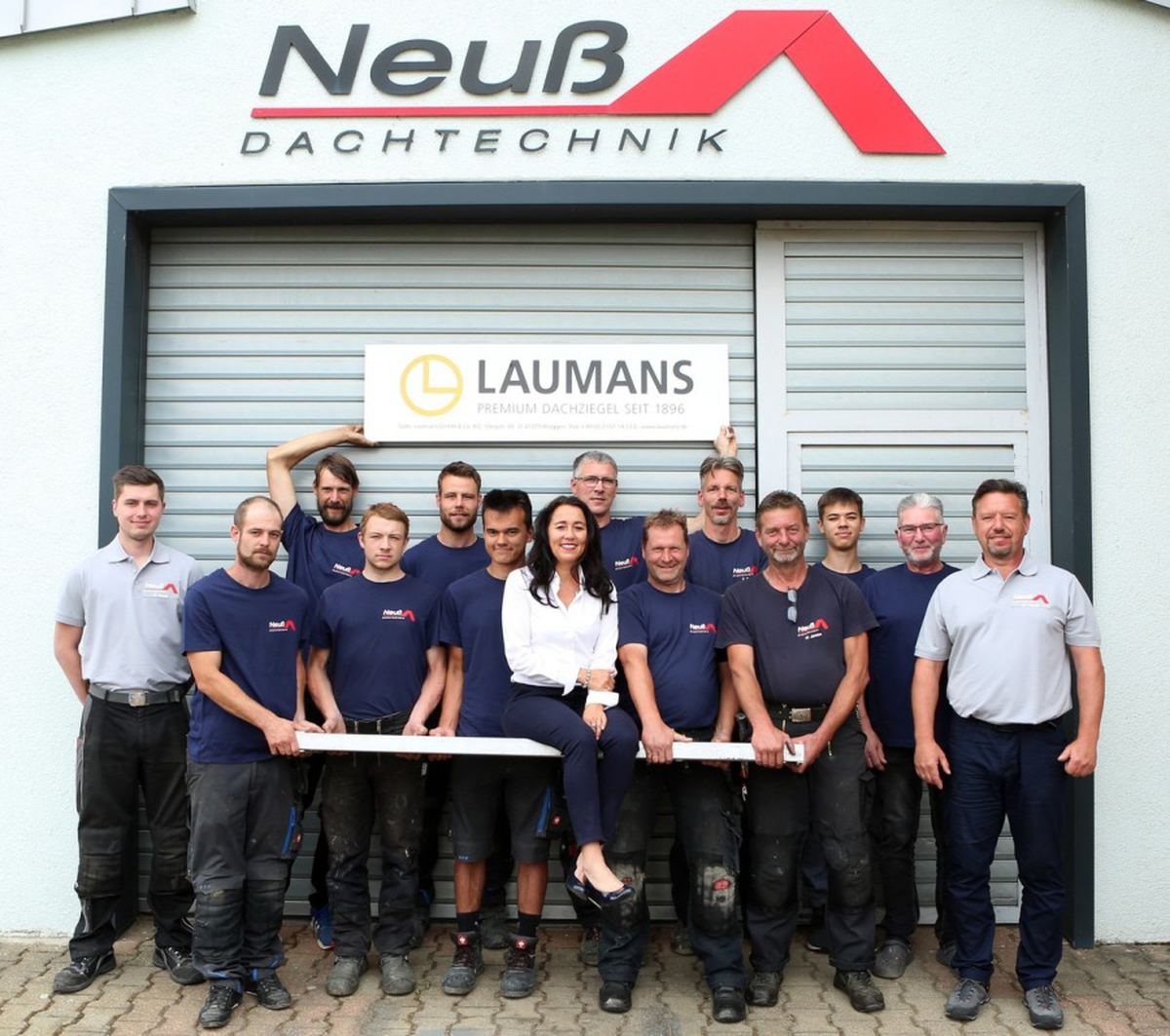 Teamfoto von Neuß Dachtechnik: 14 Personen in Firmenkleidung vor dem Tor des Betriebsgebäudes mit Neuß-Schild