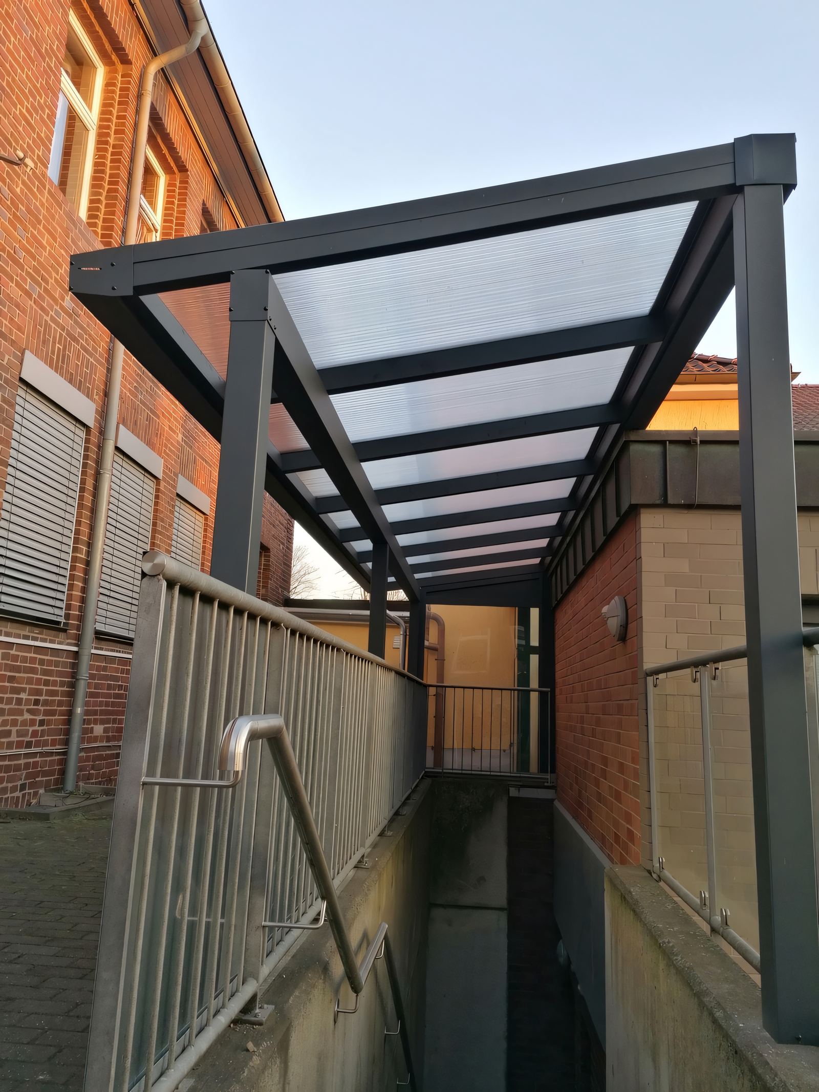 Überdachung mit anthrazitfarbenem Metallrahmen und transparenter Eindeckung über einem Kellerabgang mit Edelstahlgeländer zwischen Klinkerwänden