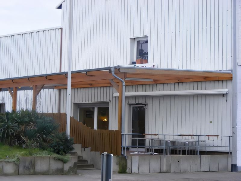 Holz-Vordach mit Metallrinne an einer weißen Gewerbehalle, darunter Eingang und Sitzplatz